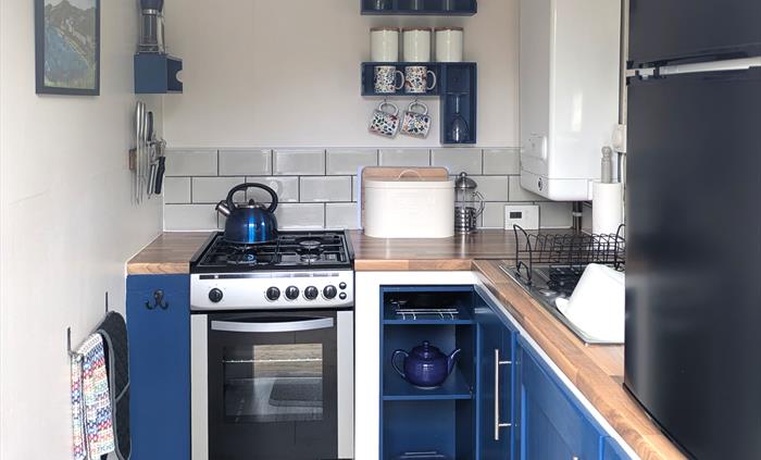 Blue cupboards and shelves. Gas cooker with hob top kettle, boiler, sink, and counter top fridge.